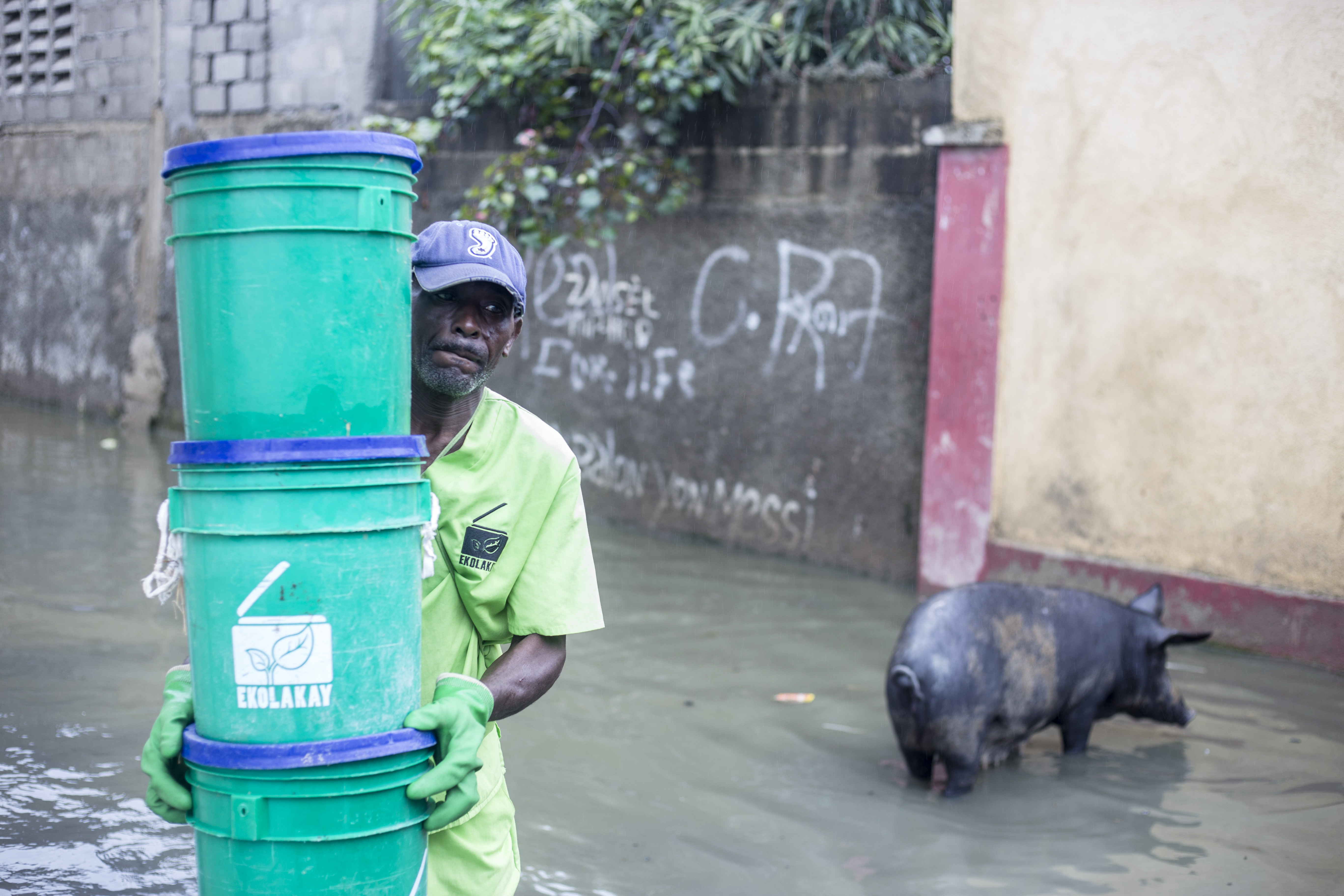 Despite flooded streets, an EkoLakay worker picks up full buckets to take for composting. Photo: Caleb Alcenat/Labelimage for Project #WasteNot (Creative Commons).